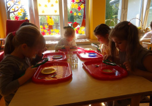 Dzieci siedzą przy stoliku i oglądają przez lupę suche drożdże wysypane na talerzykach.